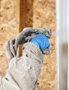 Worker in protective gear spraying foam insulation onto a wall inside a building under construction.