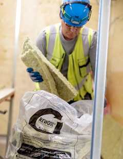 En man i blå hjälm och gul väst lägger ned stenullsavfall i en REWOOL-plastsäck.