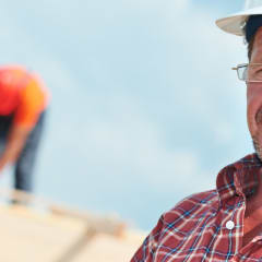 Close up of man wearing a white hard hat, adjusting glasses on his face, with blurred roofers on top of roof in background.