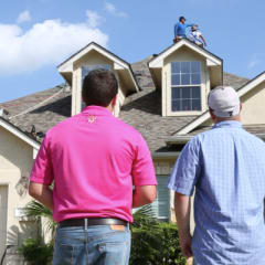 Photo of a contractor and homeowner looking at a house while roofers are working on installing shingles