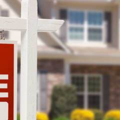 Photo of a red "For Sale" sign in the foreground, with a two story home in the background