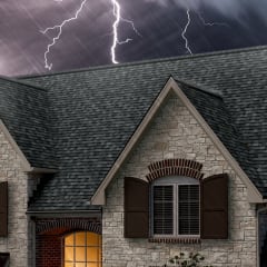 Photo of the top of a roof with gray shingles set against a dark, stormy sky with lightning