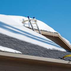 Grey roof with snow blanketed across the surface with a ladder extended up to the rooftop.