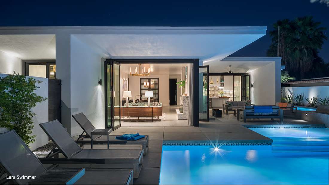 Luxury modern home with white stucco exterior, folding glass doors opening to a poolside terrace, illuminated swimming pool, and outdoor lounge chairs under night sky.