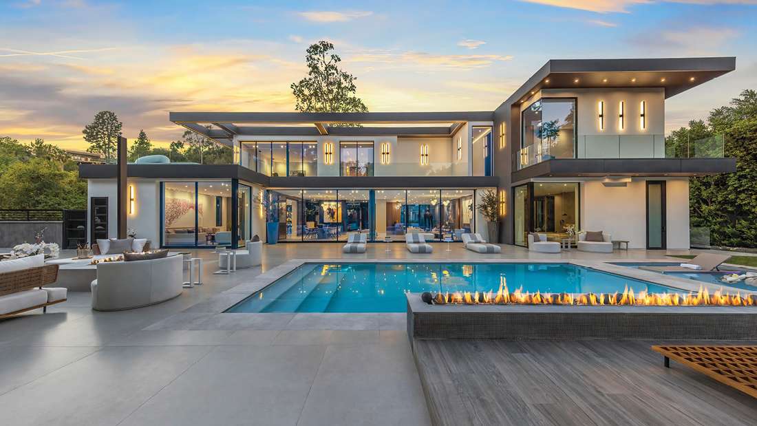 Luxury modern two-story home with floor-to-ceiling glass walls, sleek flat roof design, outdoor lounge seating, and large swimming pool with fire feature at sunset.
