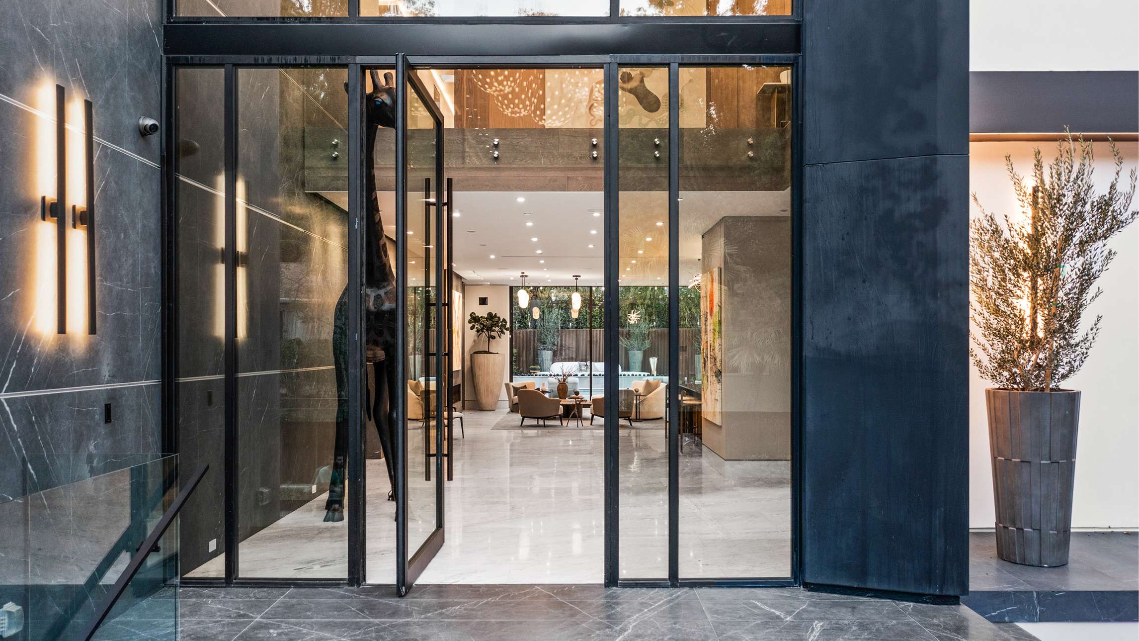 Luxury home entryway with double glass pivot doors framed in black metal, dark marble walls with vertical light fixtures, and view into open-concept living area featuring modern furniture and large potted plant.