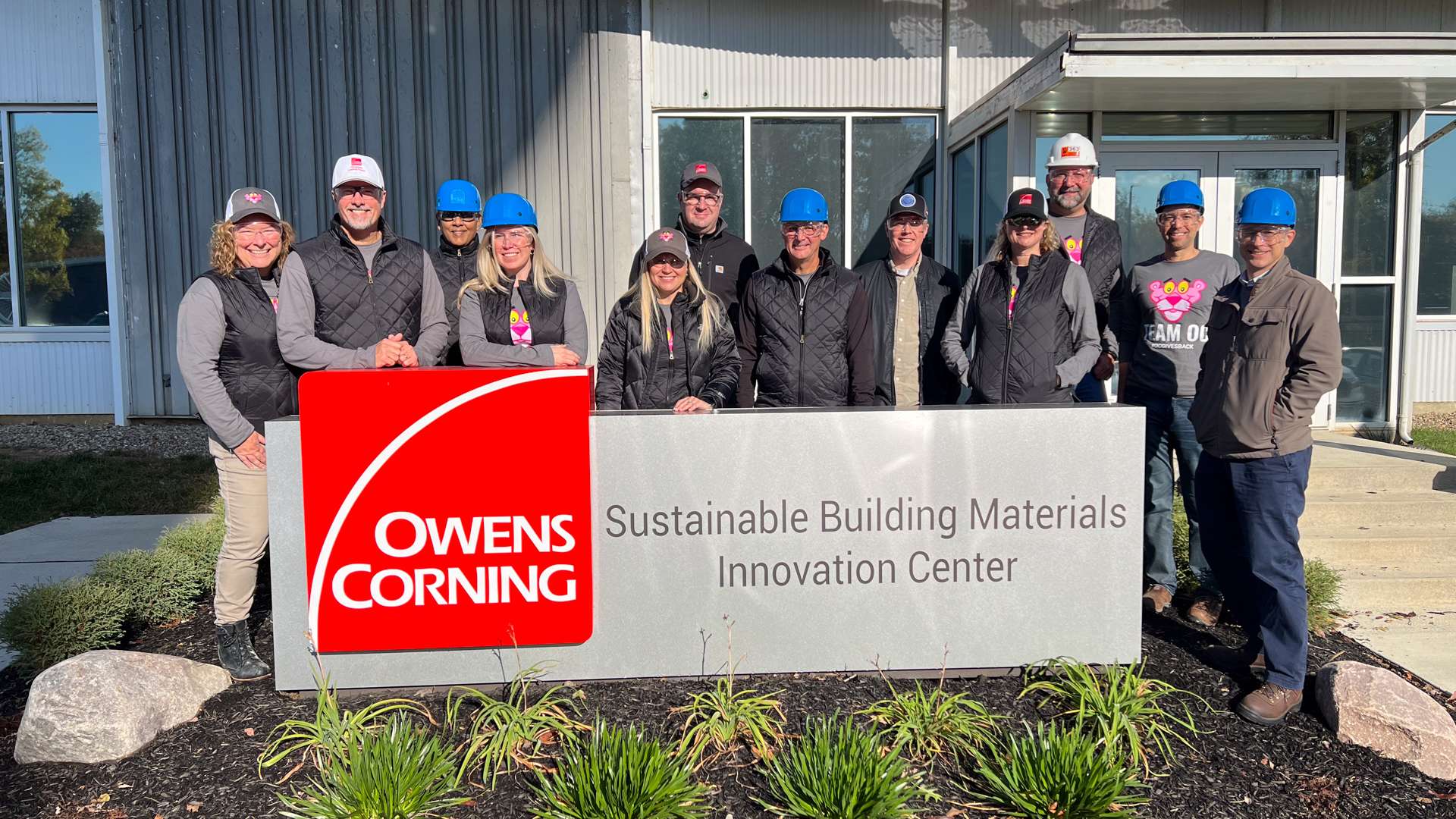Group of people standing behind a large Owens Corning sign that reads “Sustainable Building Materials Innovation Center,” in front of a modern building with metal siding and glass doors, with landscaped greenery in the foreground.