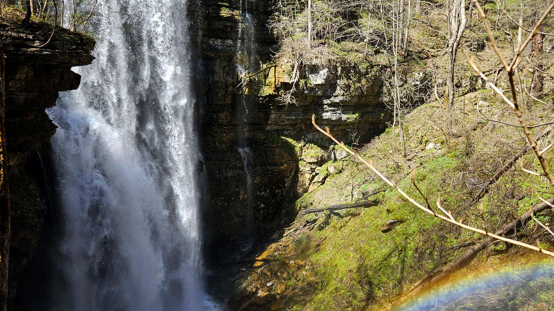 Tall waterfall cascading over rocky cliff with green moss and a rainbow at the base in a forest setting.