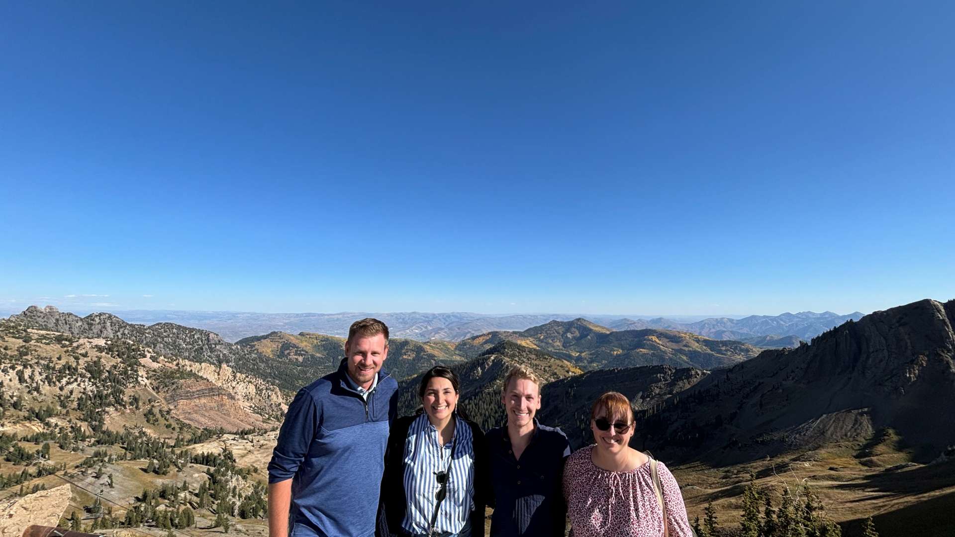 Four people standing at a mountain overlook with panoramic views of rugged peaks and valleys under a clear blue sky.