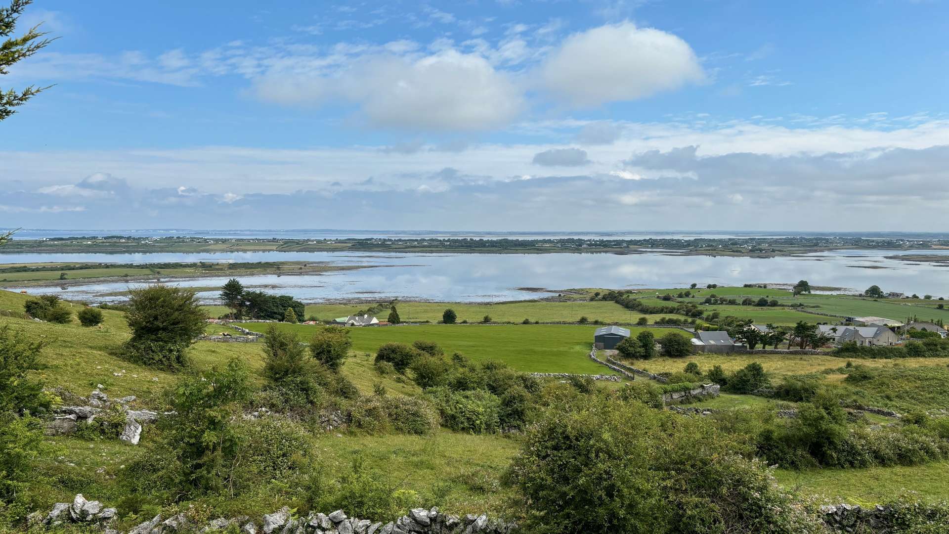 Green fields with stone walls and scattered trees overlooking a calm lake under a partly cloudy blue sky.