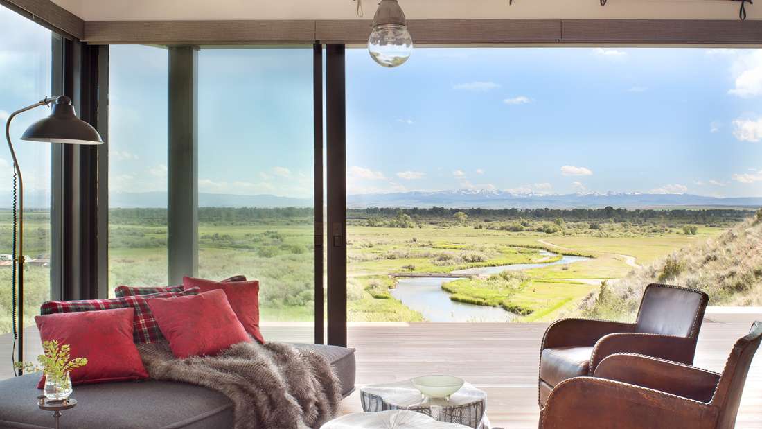 Cozy modern living space with floor-to-ceiling glass doors offering panoramic views of a winding river and grassy plains, featuring leather armchairs, plaid throw pillows, and a gray chaise lounge.
