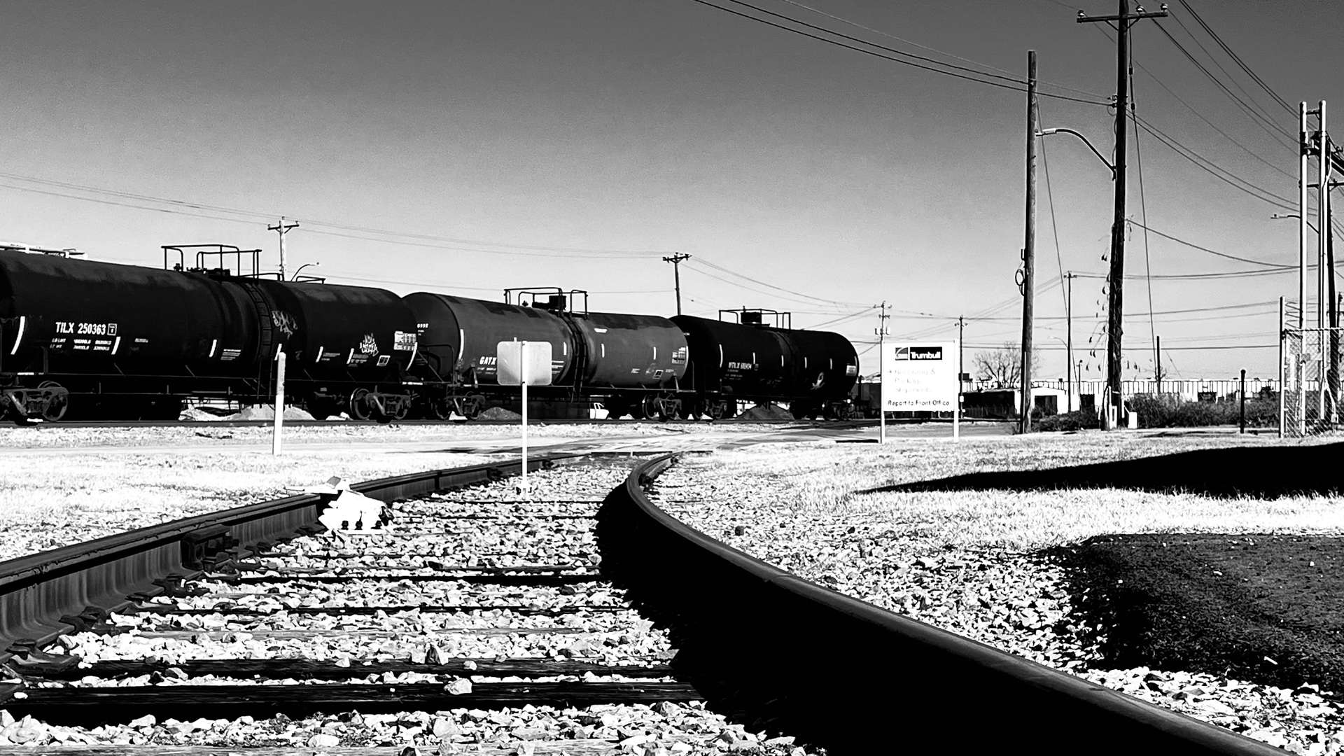 Black and white photo of train tank cars on railroad tracks with utility poles and industrial structures in the background.