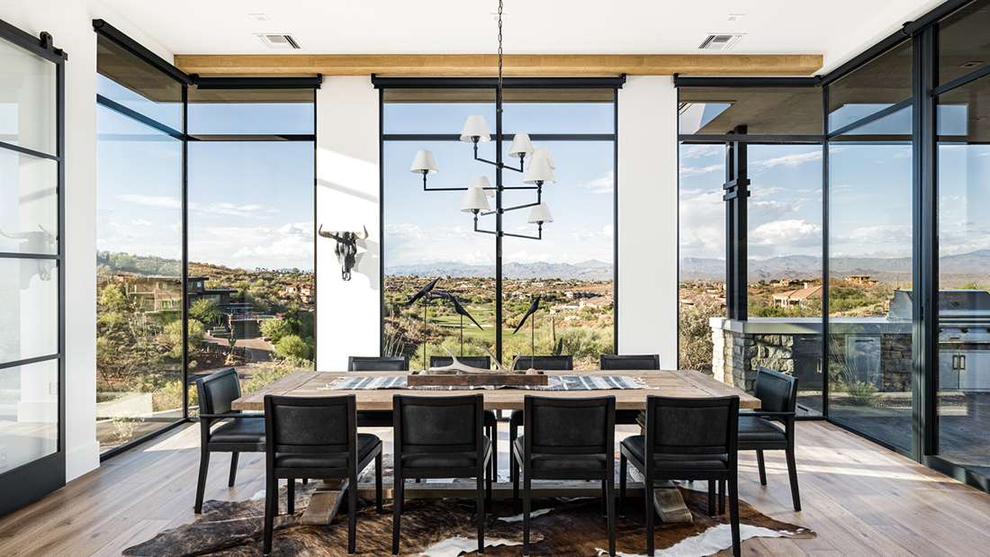 Modern dining room with large wooden table seating eight black chairs, floor-to-ceiling glass walls offering panoramic desert and mountain views, contemporary chandelier, and cowhide rug on light wood flooring.