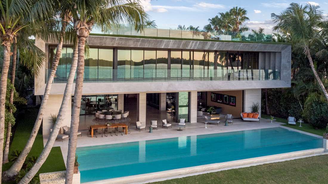 Luxury modern waterfront home with minimalist concrete exterior, expansive floor-to-ceiling glass walls, wraparound balcony with glass railing, and outdoor living space featuring lounge seating and infinity-edge pool surrounded by palm trees.