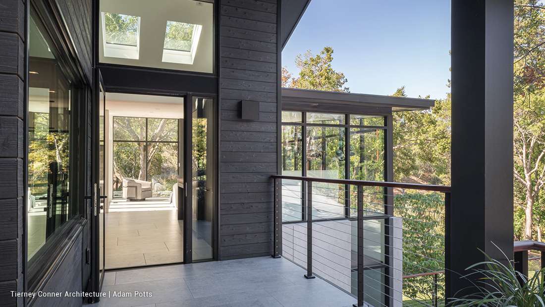 Modern home exterior with dark wood siding, floor-to-ceiling glass doors, and balcony railing featuring metal cables, overlooking lush greenery and trees.