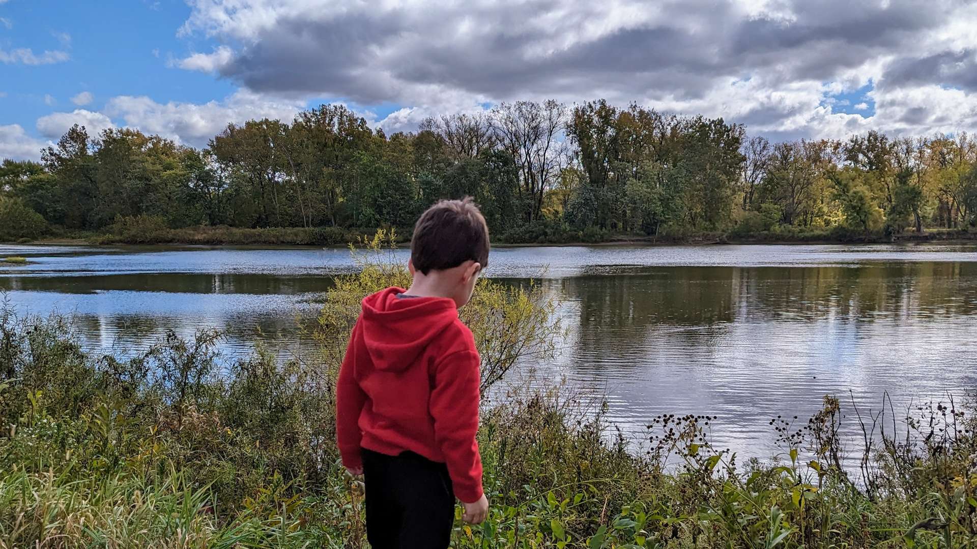 Child wearing a red hoodie standing near the edge of a calm river, surrounded by tall grass and vegetation, with a backdrop of autumn trees and a partly cloudy sky.