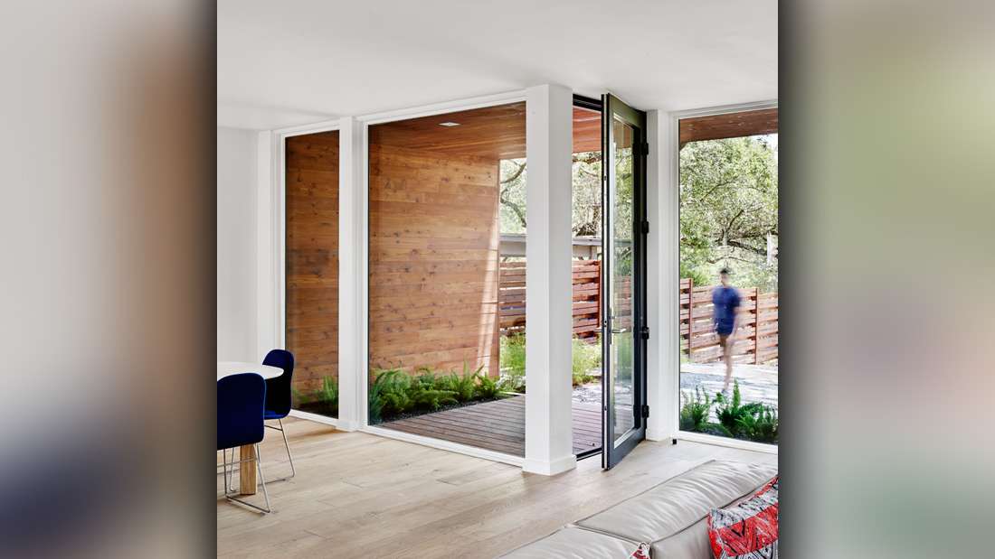 Modern living space with folding glass door opening to outdoor patio, floor-to-ceiling windows framed in white, natural wood siding on exterior walls, light wood flooring, and minimalist furniture with colorful accent pillows.