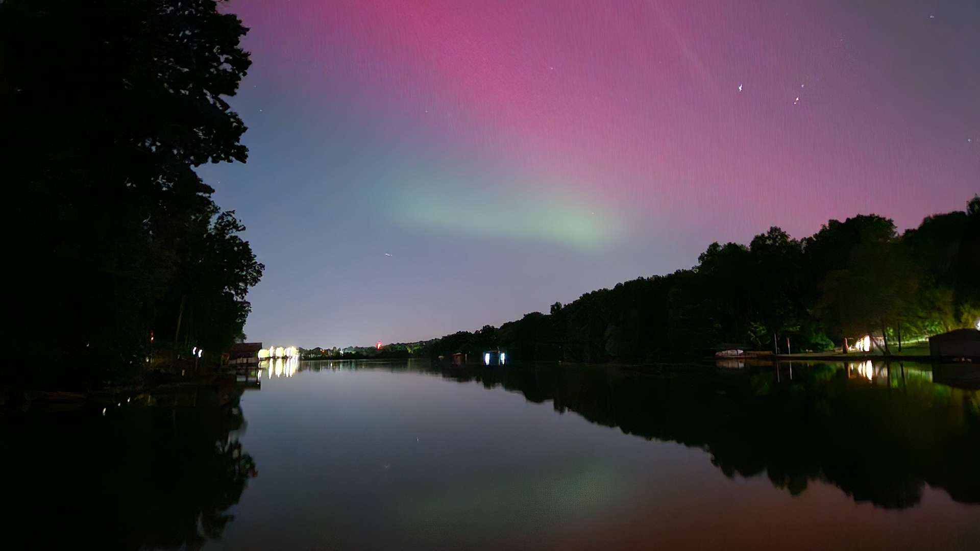   Northern lights with pink and green hues reflecting on a calm lake surrounded by trees at night.