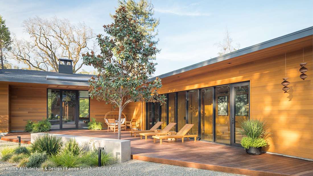 Modern single-story home with horizontal wood siding, black-framed glass doors, spacious wooden deck featuring lounge chairs and dining area, and minimalist landscaping with potted plants and a central tree.