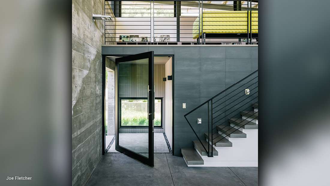 Modern entryway with pivot glass door framed in black metal, gray tiled walls, minimalist staircase with black railing, and upper loft featuring metal balustrade and bright yellow bedding.