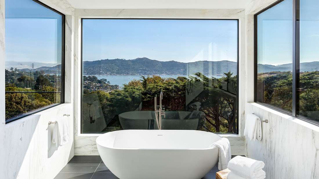 Luxury bathroom with freestanding white oval bathtub positioned in front of a large picture window offering panoramic views of a bay, surrounding hills, and lush greenery, framed by marble walls and gray tile flooring.