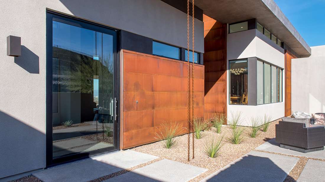 Contemporary desert home exterior with smooth stucco walls, rust-colored metal panels, large black-framed glass door, and minimalist xeriscape landscaping featuring gravel and drought-tolerant plants.