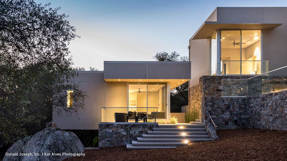 Contemporary hillside home with stone foundation, glass railings, large windows, and illuminated entry steps at dusk.