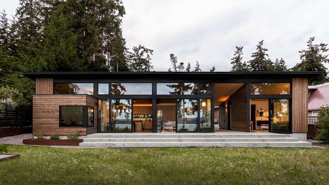 Modern single-story home with flat roof, horizontal wood siding, large floor-to-ceiling glass windows, and open-concept interior featuring warm wood accents surrounded by tall evergreen trees.