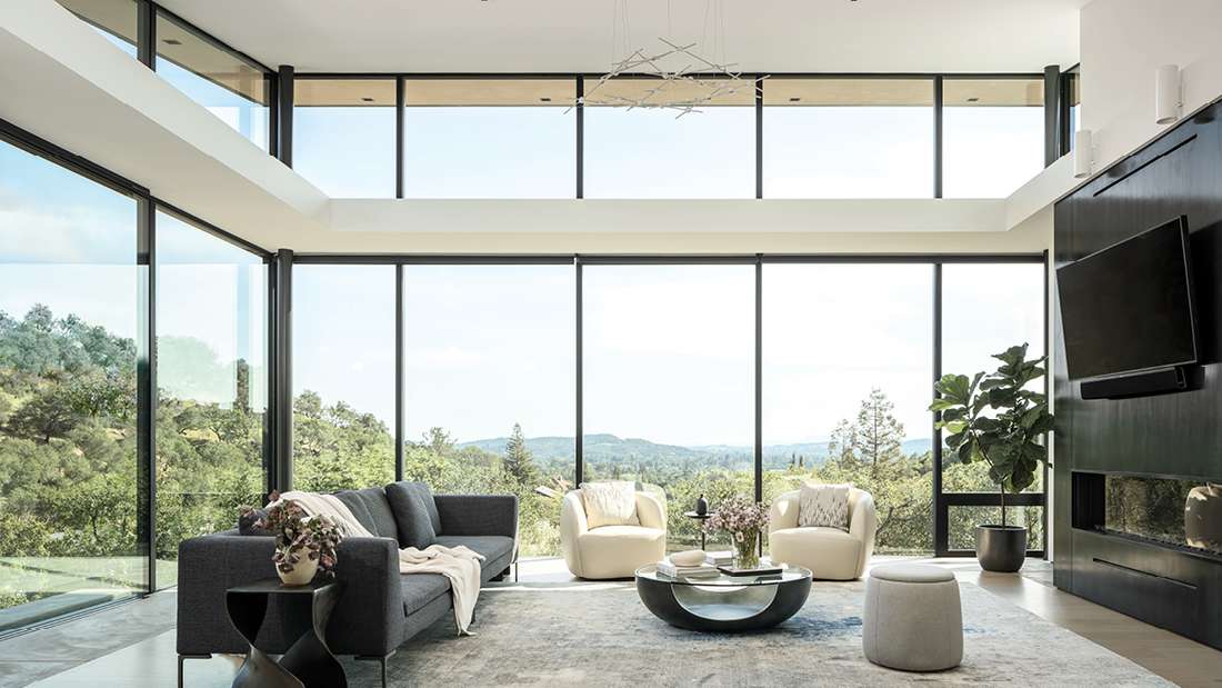 Luxury living room with double-height floor-to-ceiling glass walls offering panoramic forest views, dark gray sofa paired with two cream armchairs, round modern coffee table, and sleek black accent wall featuring mounted TV and linear fireplace.