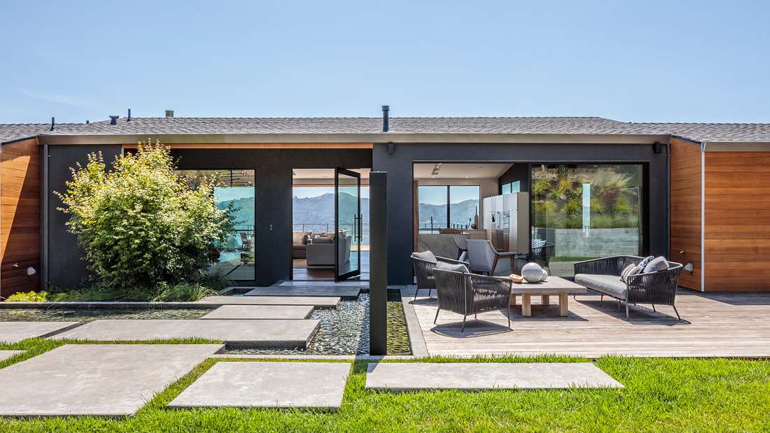 Modern single-story home exterior with black stucco walls, natural wood siding accents, large glass doors opening to outdoor seating area, and minimalist landscaping featuring concrete pavers and green lawn.