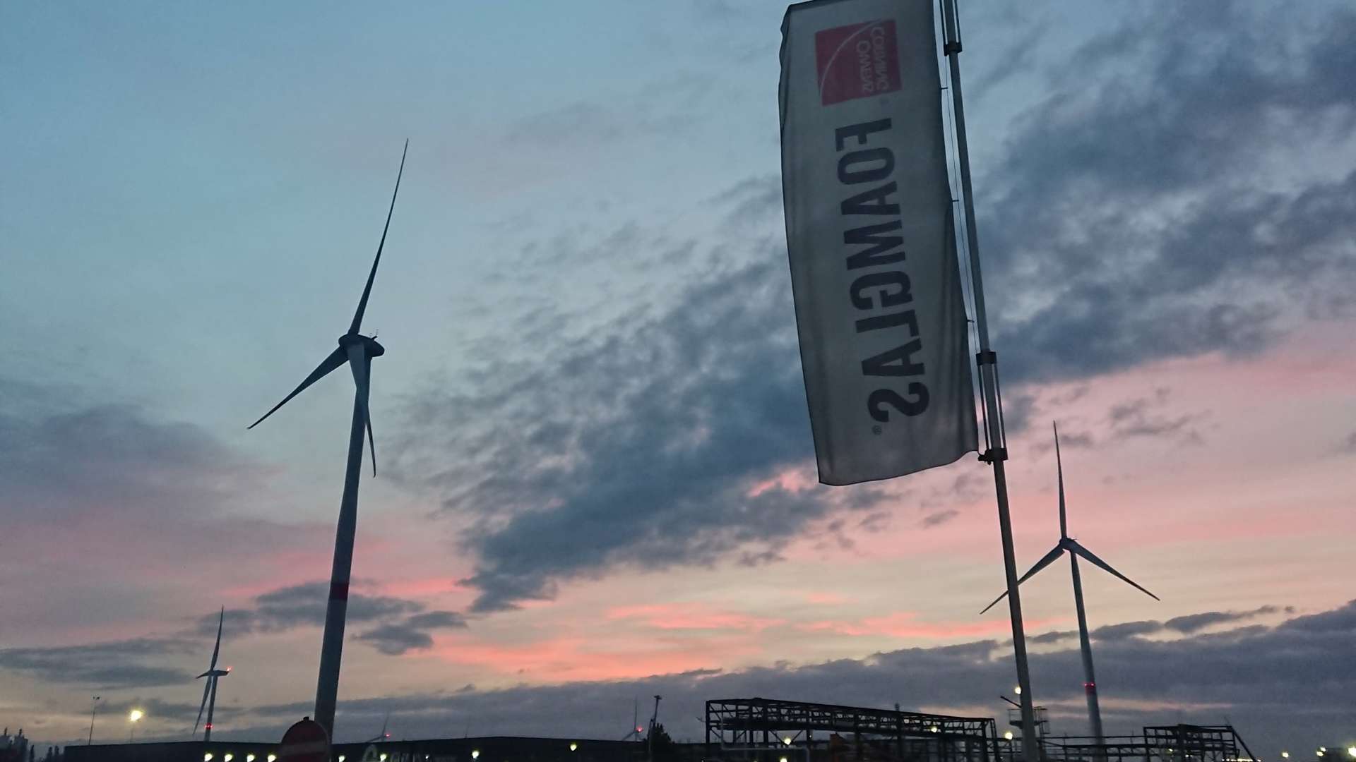 Wind turbines and a vertical flag at sunset with pink and blue clouds over an industrial site and silhouetted structures.