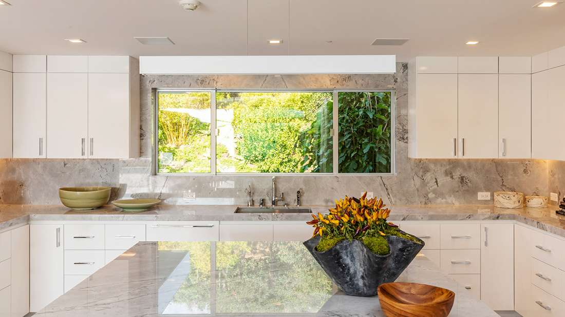 Luxury modern kitchen with white cabinetry, marble countertops and backsplash, large center island with polished stone surface, and wide window overlooking lush green garden.