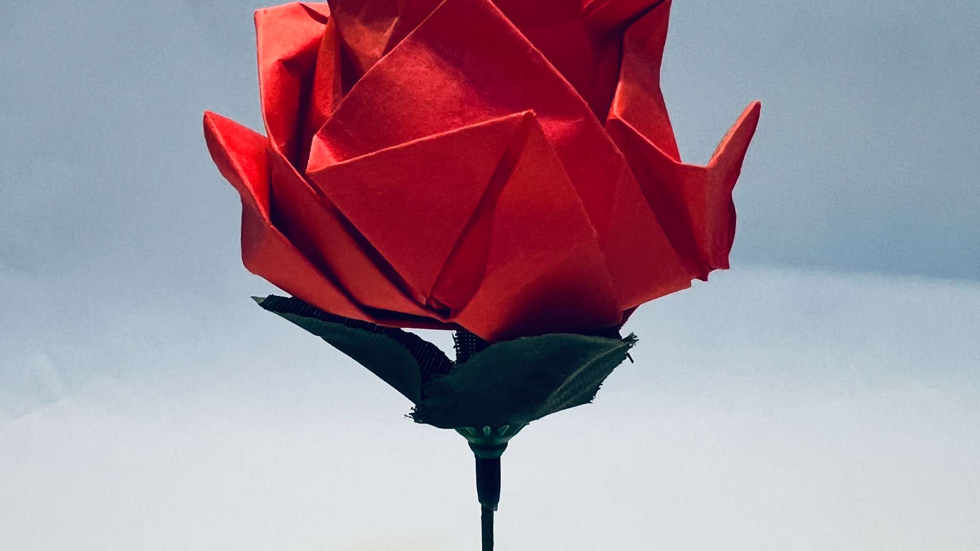   Origami red rose with green leaves on a thin stem against a soft gradient background.