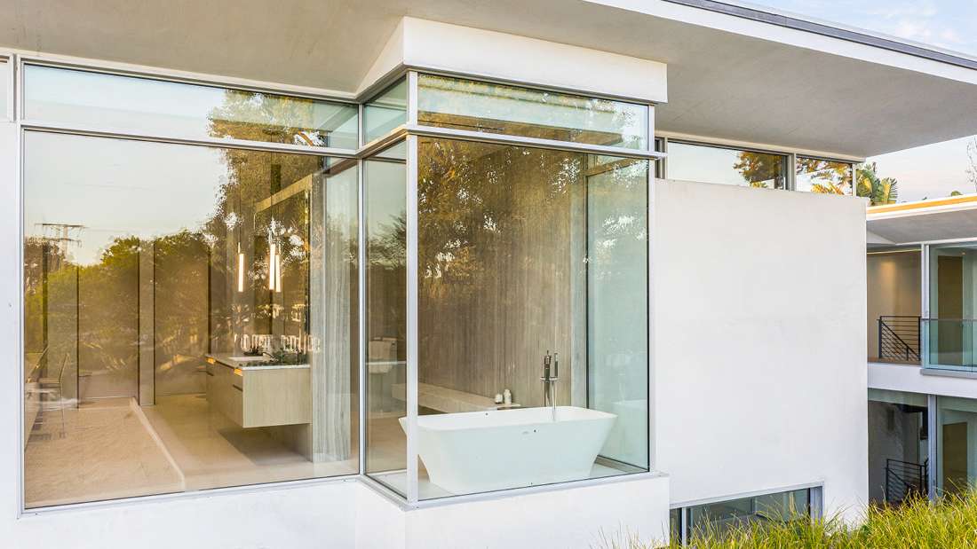 Modern bathroom with freestanding white bathtub positioned in a glass corner enclosure, floor-to-ceiling windows offering outdoor views, and floating vanity with pendant lights.