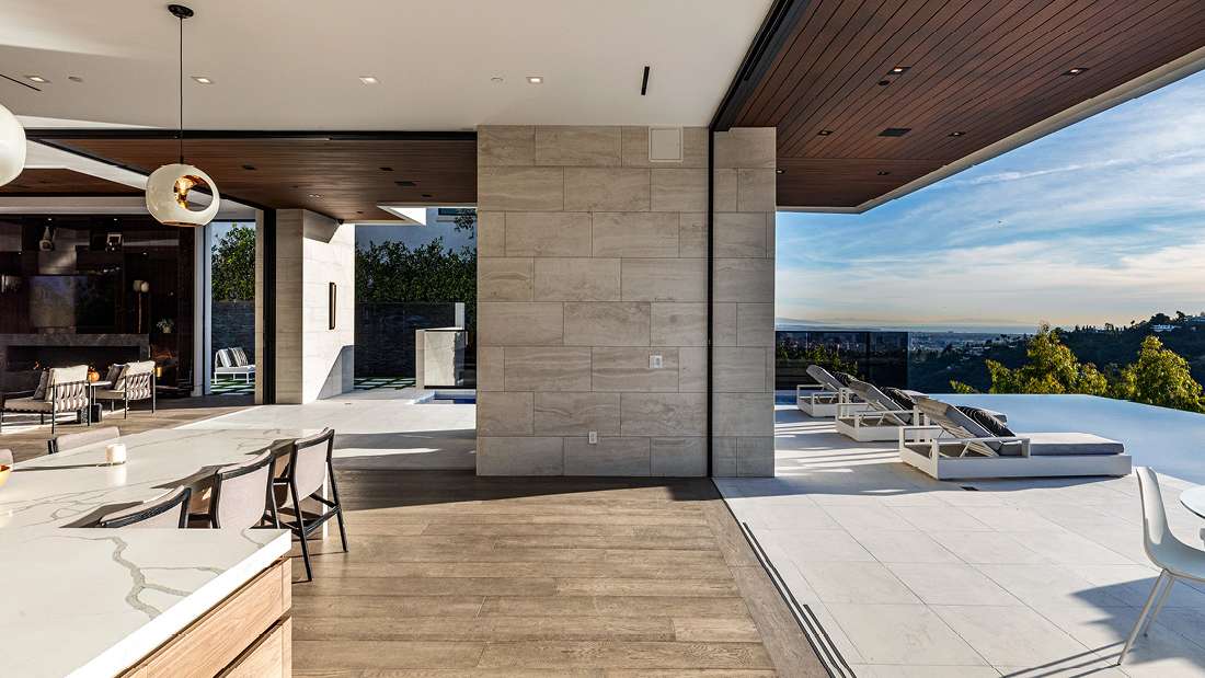 Luxury open-concept home interior with marble kitchen island, wood flooring, and sliding glass walls opening to a terrace with lounge chairs and infinity pool overlooking hillside views.