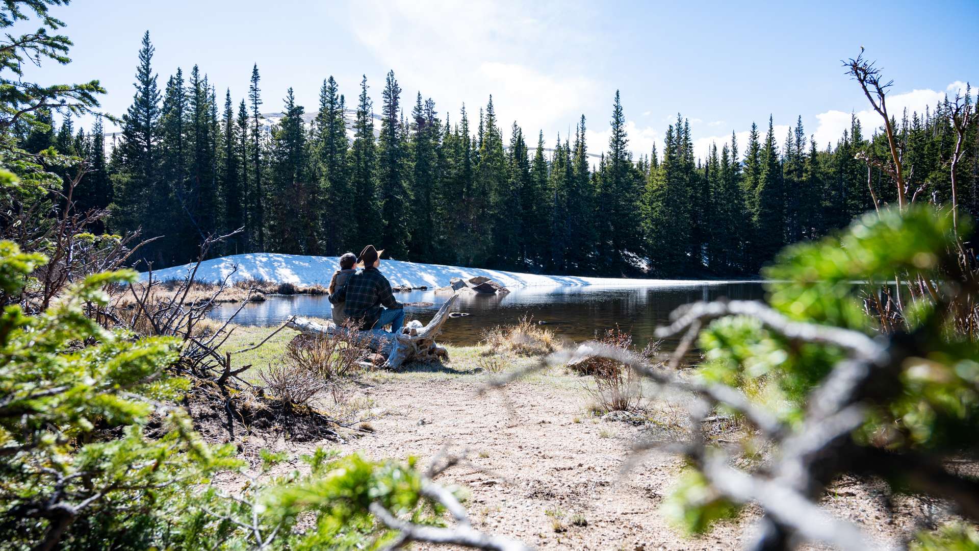   Two people sitting on a sandy lakeshore near snow patches, surrounded by evergreen trees under a bright blue sky.