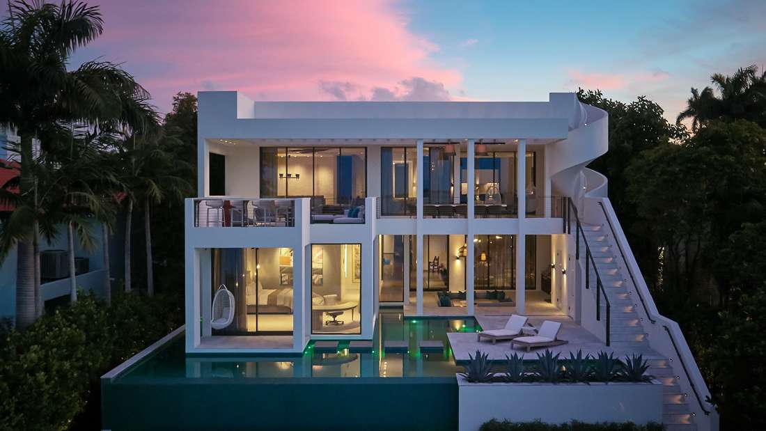 Luxury modern home with minimalist white exterior, expansive floor-to-ceiling glass walls, wraparound balcony, and infinity-edge pool illuminated at dusk, surrounded by palm trees and lush greenery.