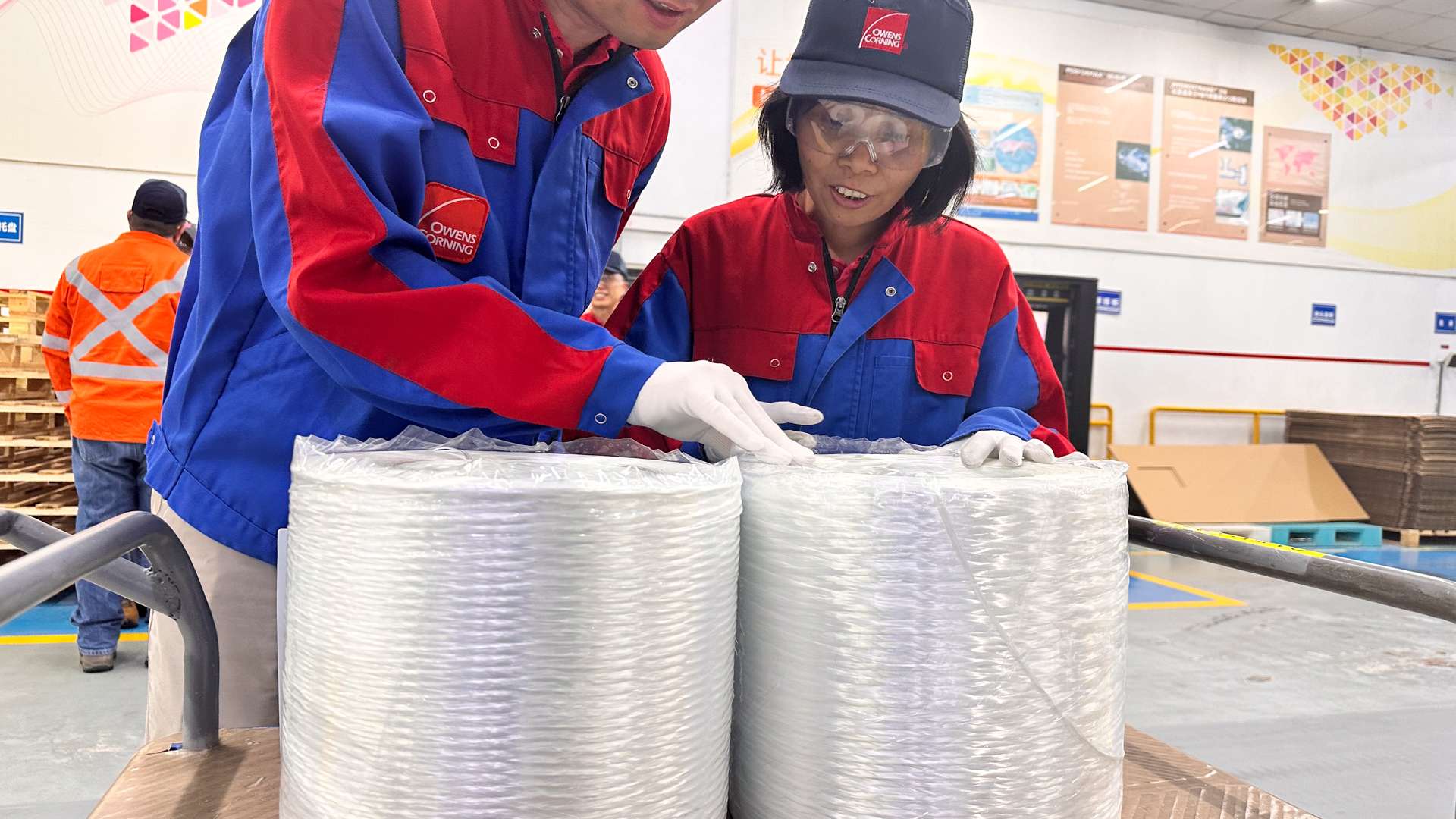 Two workers in blue and red uniforms inspecting large spools of industrial fiber on a wooden cart inside a factory setting, with safety signage and equipment visible in the background.