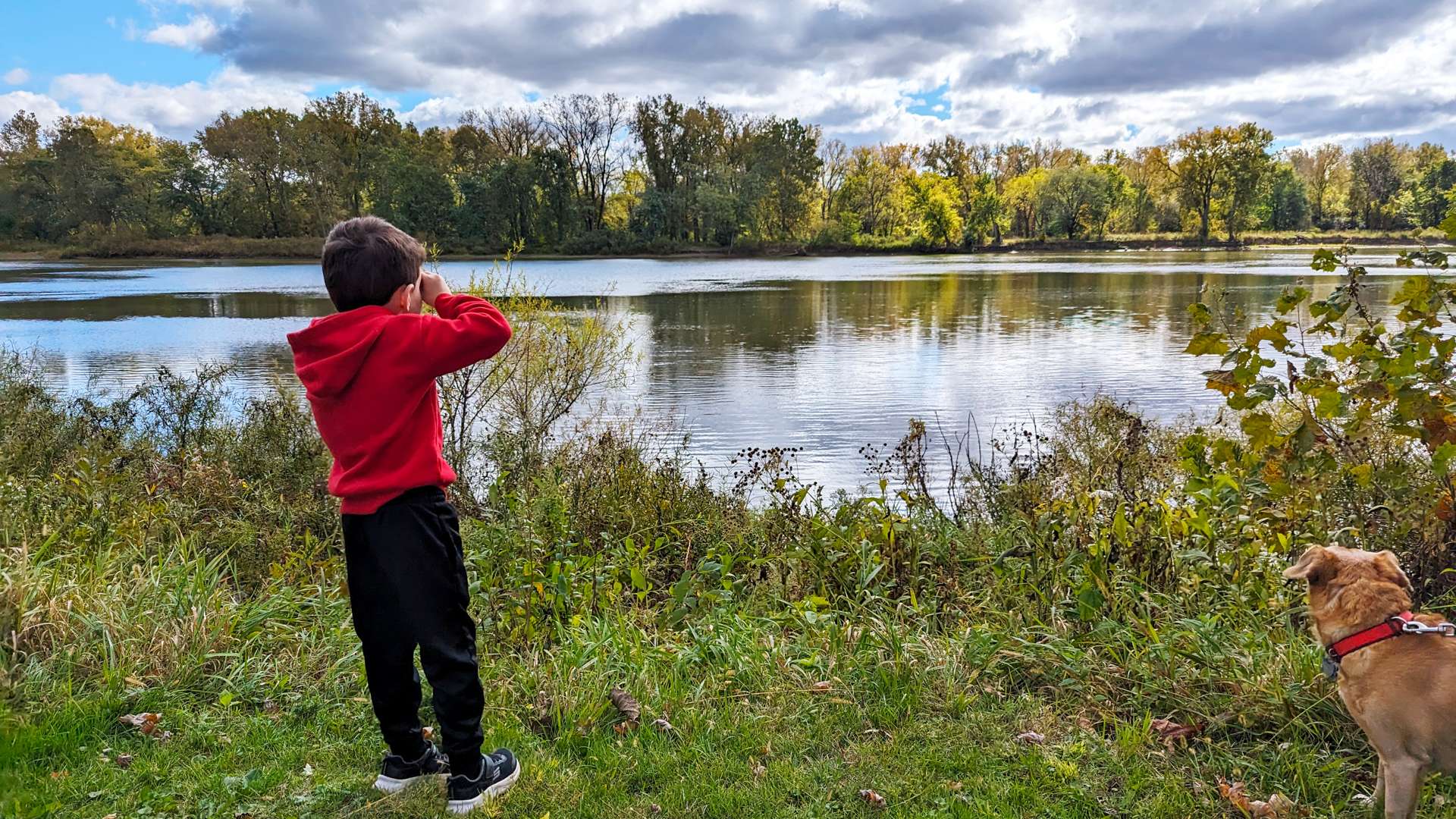 Child wearing a red hoodie looking through pretend hand binoculars near the edge of a calm river, with a tan dog on a red leash standing nearby on green grass, surrounded by autumn trees under a partly cloudy sky.