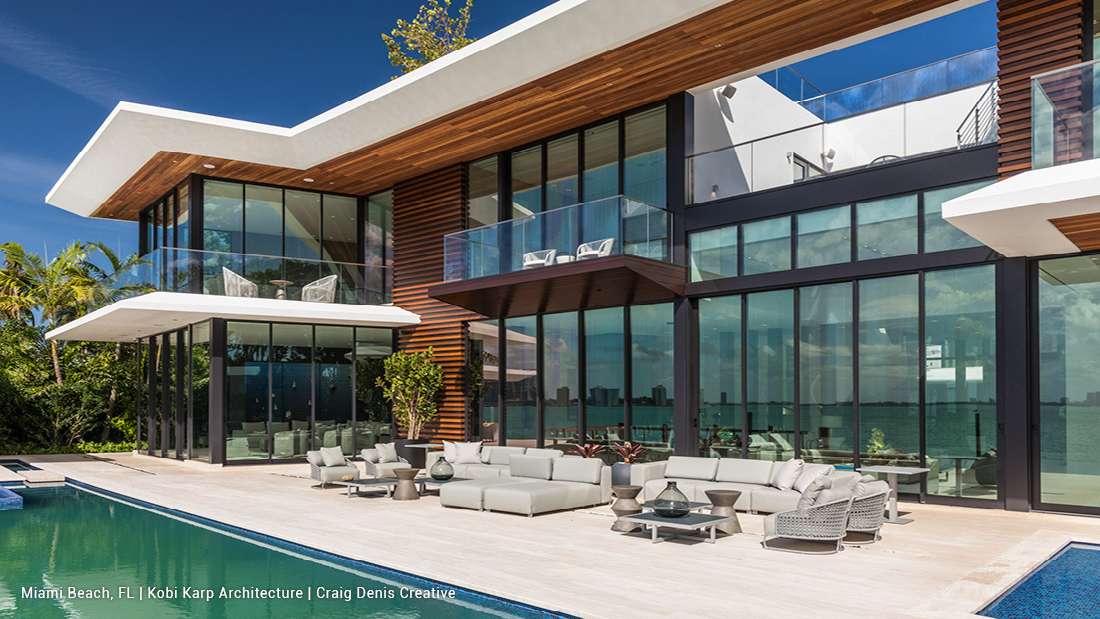 Luxury modern home in Miami Beach, Florida featuring angled rooflines, natural wood soffits, vertical wood siding accents, expansive floor-to-ceiling glass walls, and outdoor living space with lounge seating and swimming pool overlooking the Bay.