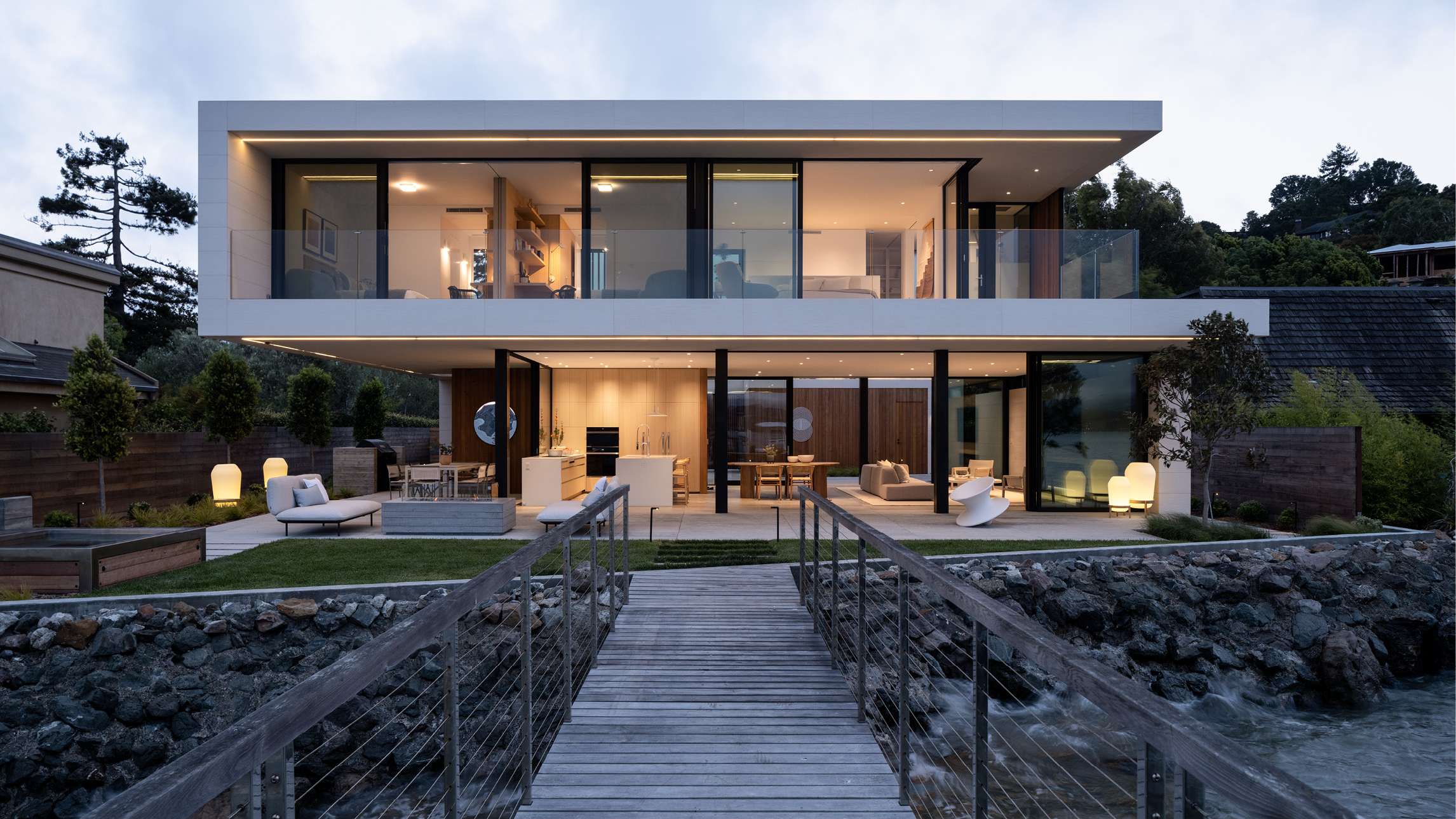 Modern two-story waterfront home with floor-to-ceiling glass walls, cantilevered upper level featuring glass balcony railings, open-concept ground floor with outdoor lounge seating, and wooden dock leading to the property.