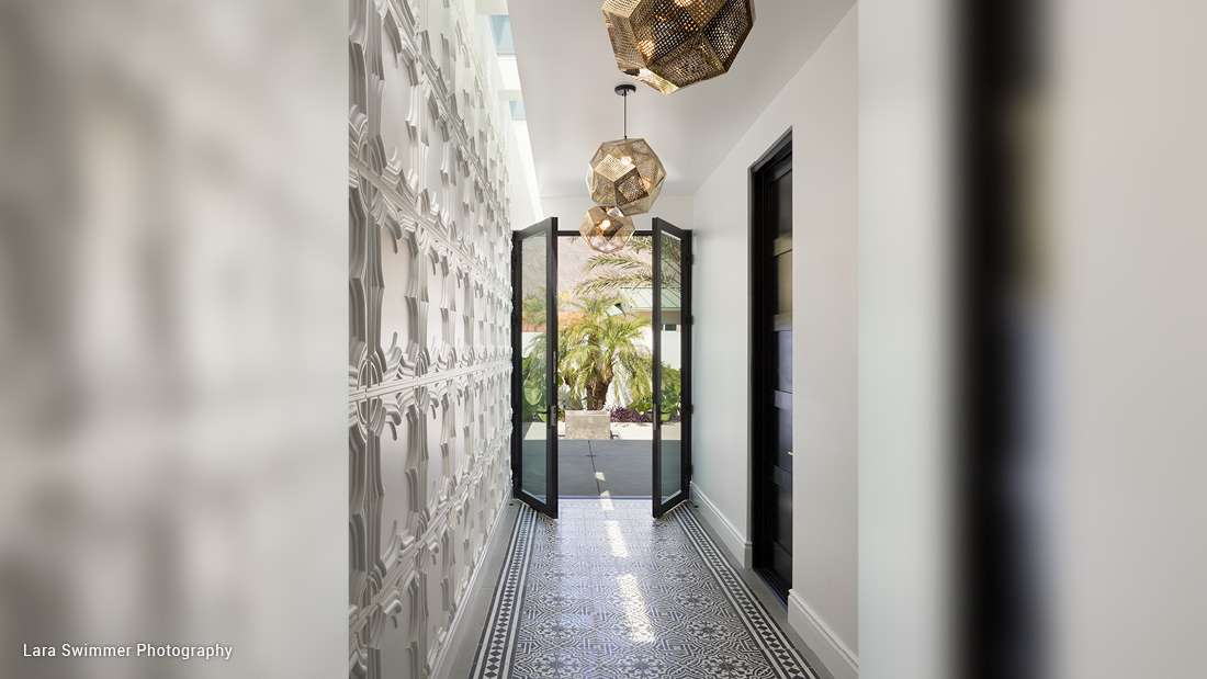 Modern hallway with decorative 3D wall panels, patterned tile flooring, geometric pendant lights, and double glass doors opening to a lush outdoor garden.