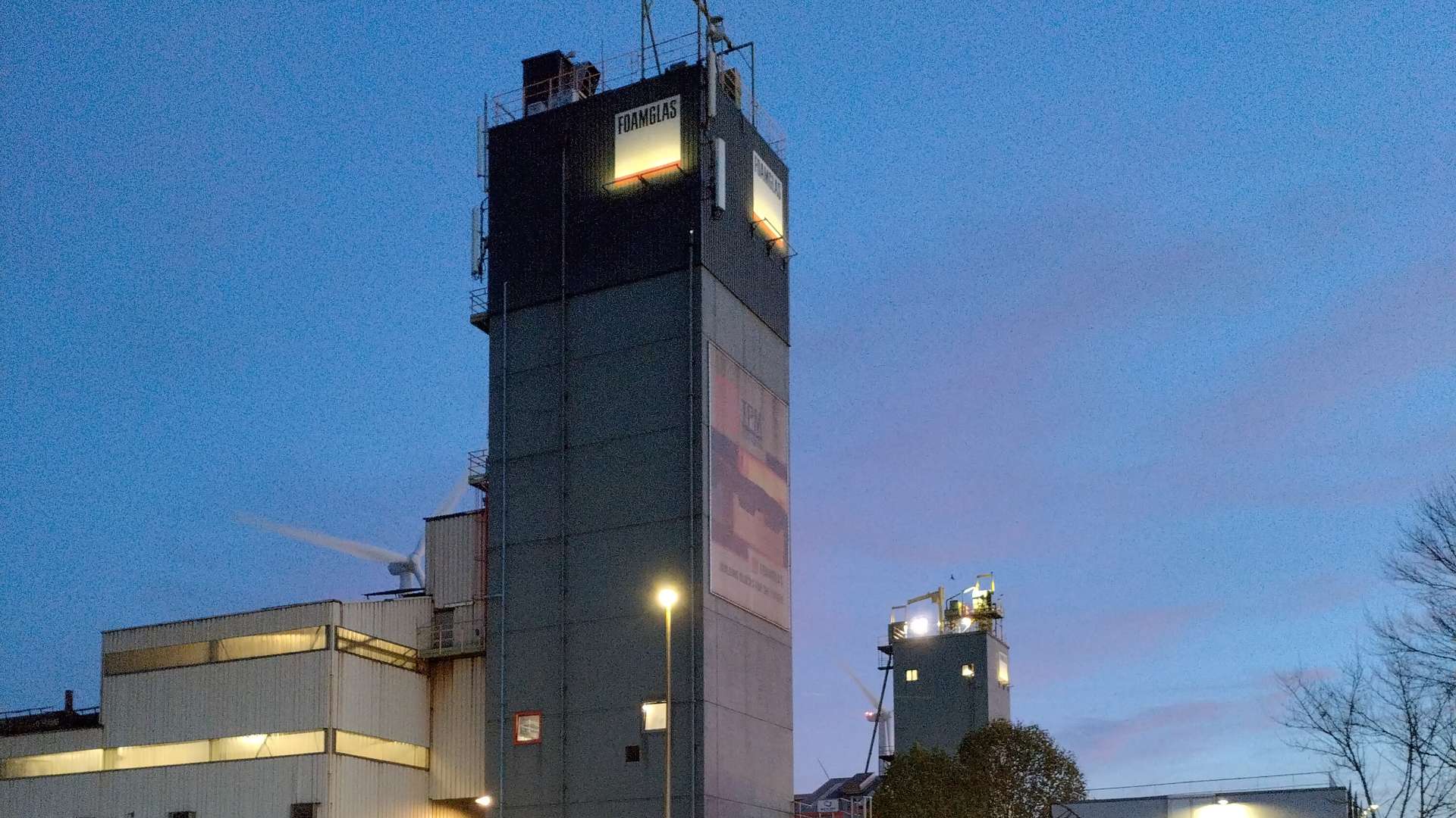 Industrial buildings with tall Foamglas-branded towers illuminated at dusk against a clear blue evening sky. 