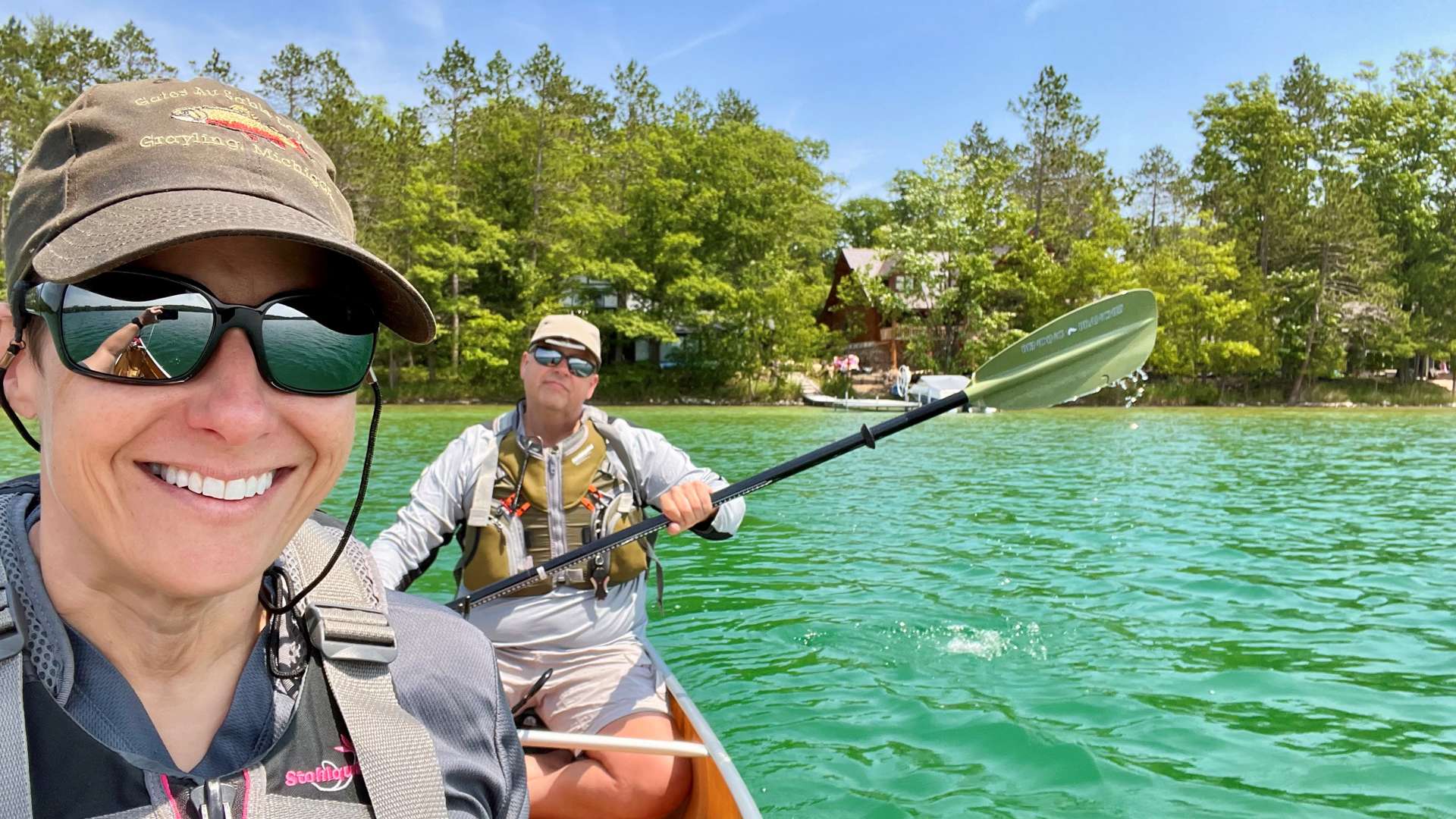 Two people paddling a canoe on clear green water near a forested shoreline with cabins visible in the background under a bright blue sky.