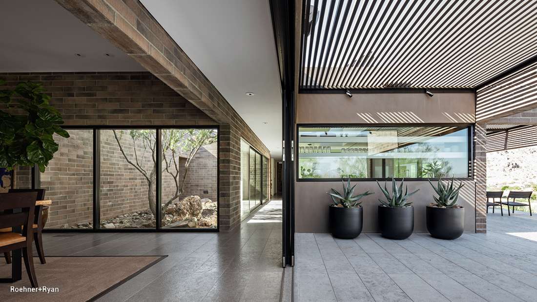 Modern courtyard with brick walls, large glass windows, and shaded patio featuring black planters with agave plants under slatted pergola.