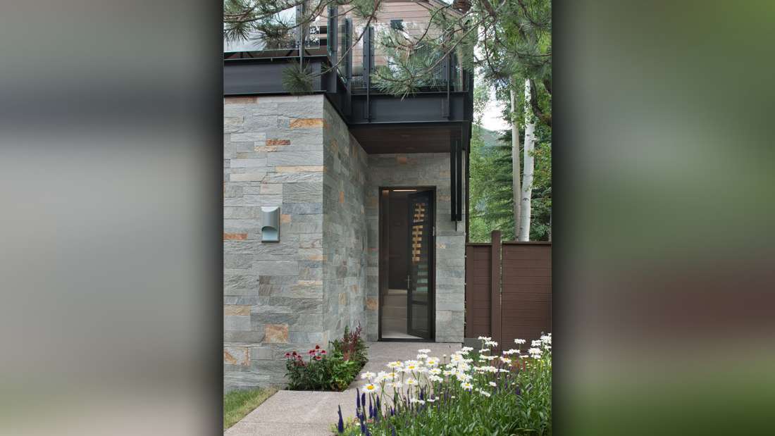 Modern home exterior with stacked stone facade, glass balcony railing, and narrow glass entry door surrounded by lush landscaping and blooming flowers.