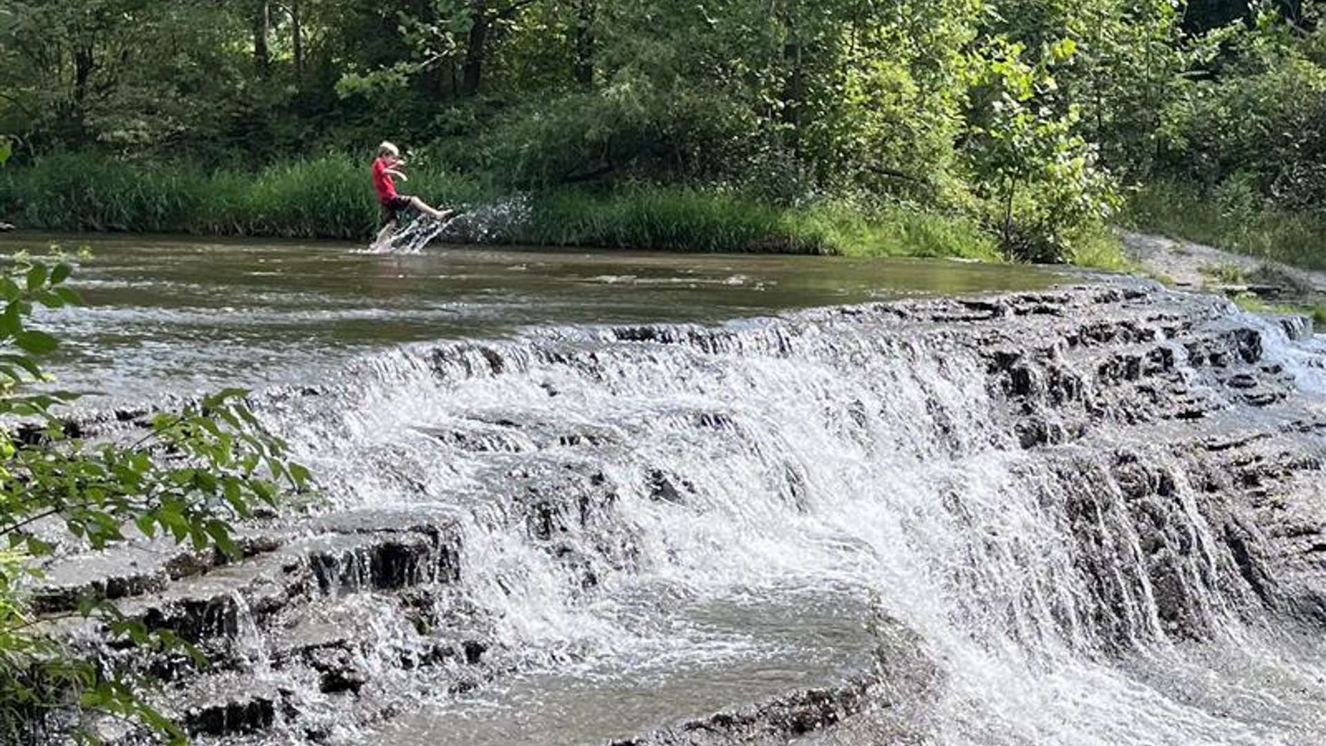 Tiered rock ledges with a small waterfall cascading into a shallow stream, surrounded by dense green forest, with a person in a red shirt standing in the water near the top of the falls.