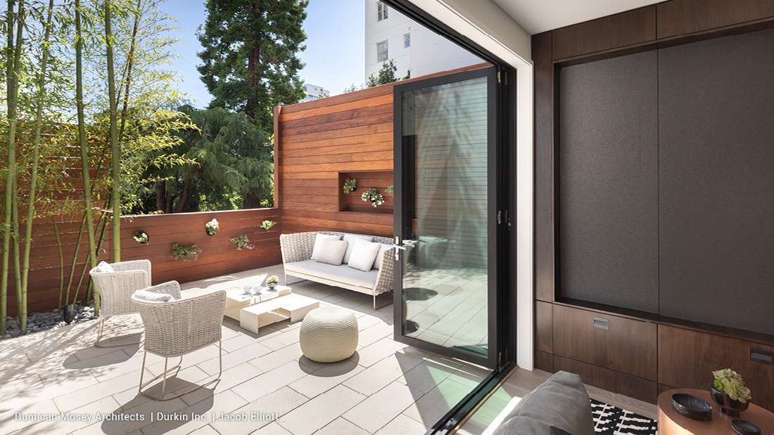 Modern outdoor patio with folding glass doors, warm wood privacy wall featuring built-in planters, wicker lounge chairs, cushioned sofa, and minimalist coffee table surrounded by bamboo and greenery.