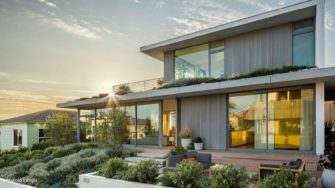 Modern two-story home with vertical wood siding, large glass windows, rooftop greenery, and landscaped front yard featuring succulents and outdoor seating at sunset.