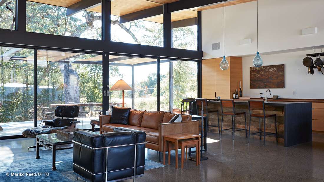 Modern open-concept living space with floor-to-ceiling glass walls, exposed wood ceiling, leather seating, and stylish kitchen with pendant lighting.